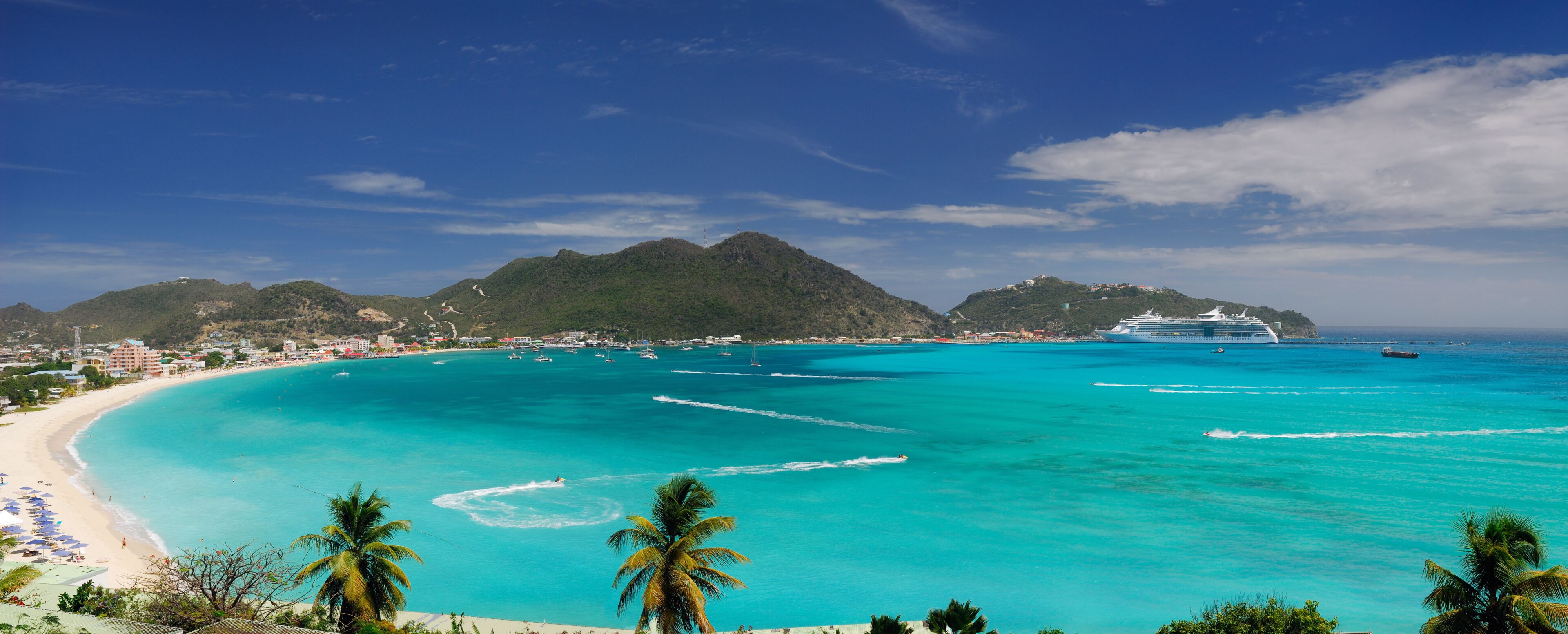 A turquoise beach with a cruise ship, palm trees, and a mountainous backdrop.