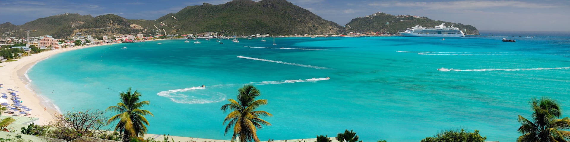 A turquoise beach with a cruise ship, palm trees, and a mountainous backdrop.
