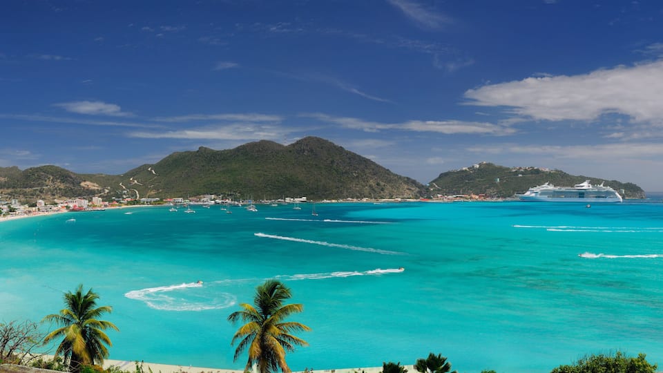 A turquoise beach with a cruise ship, palm trees, and a mountainous backdrop.