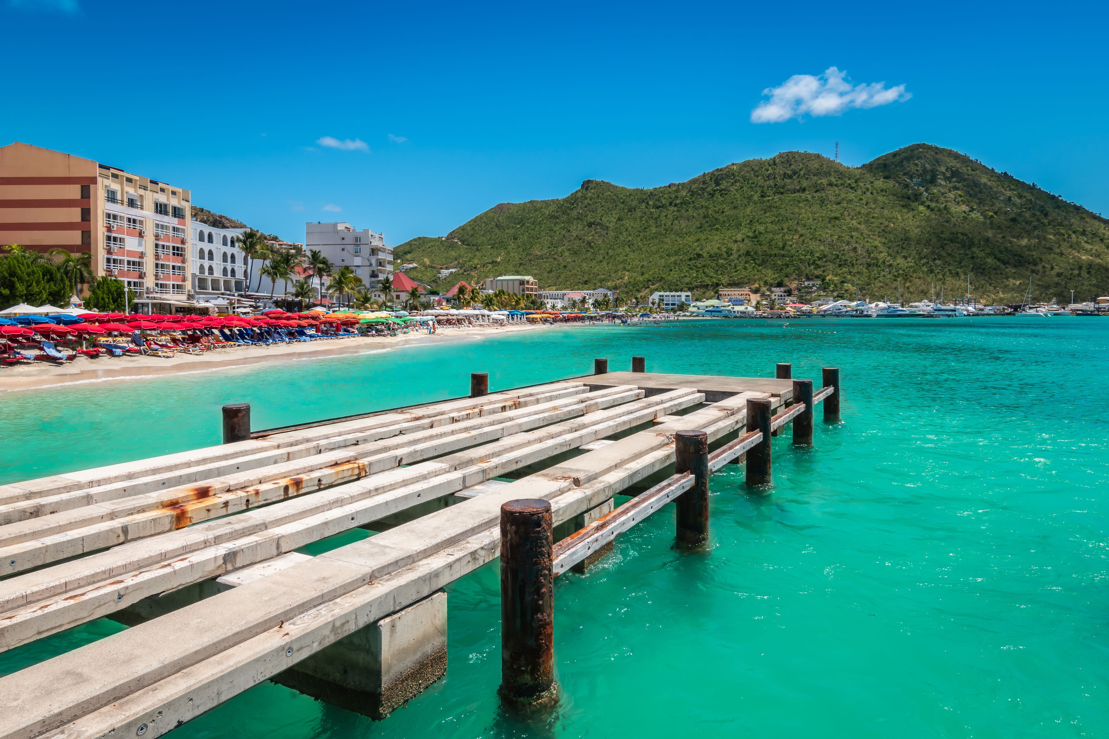 A wooden pier extending into turquoise waters, a beach with colorful umbrellas, and a mountainous backdrop.