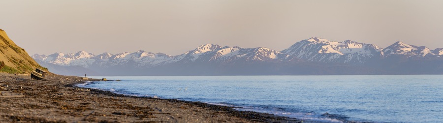 A rocky beach with a fishing boat, a person standing on the shore, and snow-capped mountains in the background.