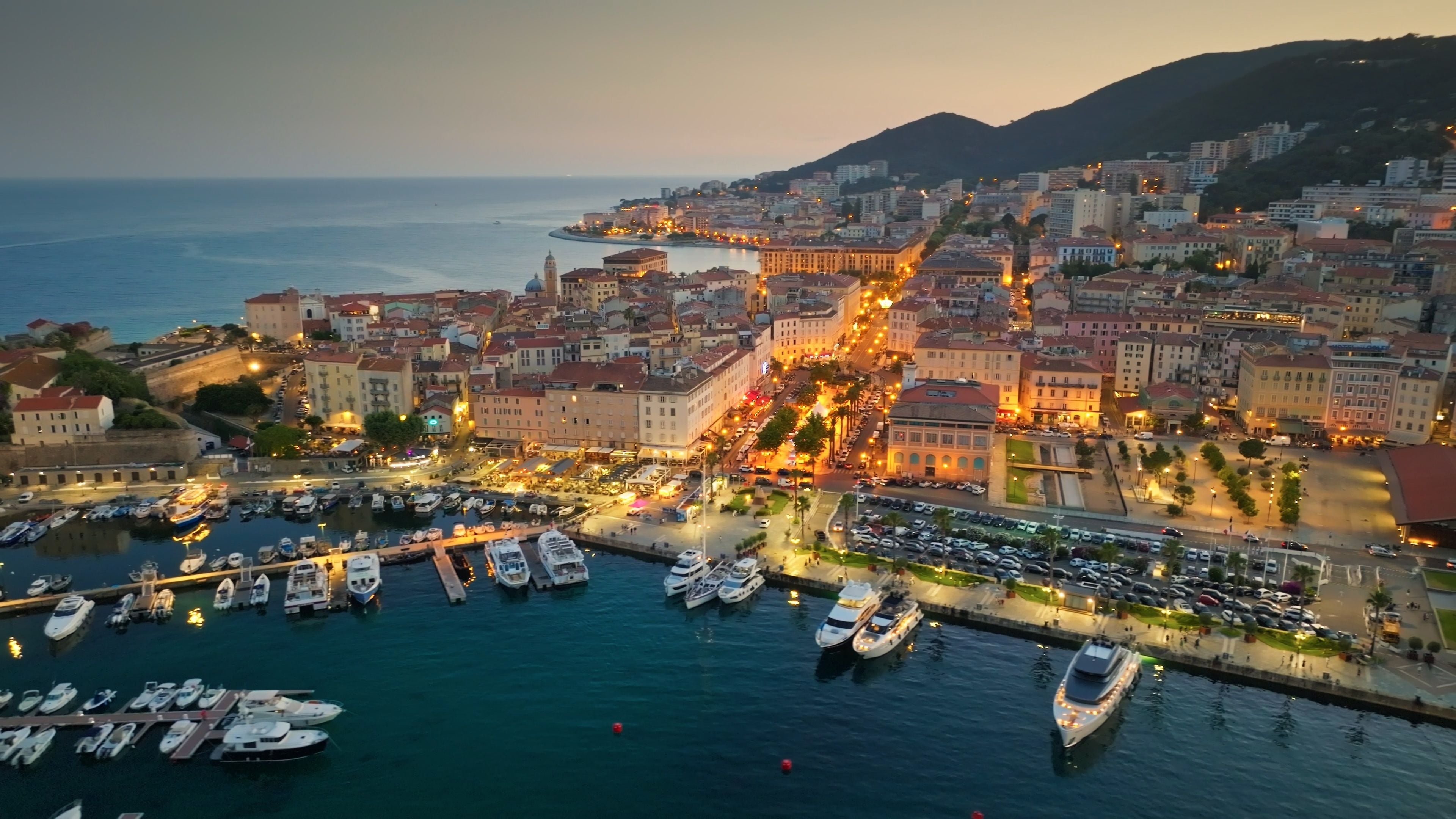 A coastal cityscape at dusk with a marina filled with boats, historic buildings, and a hilly backdrop.