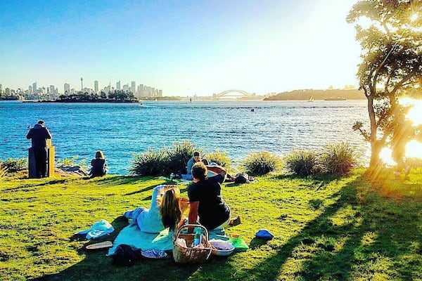 Uma área de piquenique à beira do rio, com pessoas relaxando, um horizonte urbano ao fundo e uma ponte sobre as águas.