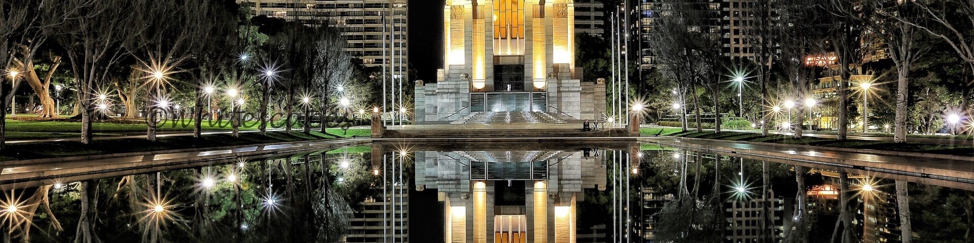 Paisagem urbana noturna com um edifício iluminado em destaque, refletido na água, cercado por arranha-céus modernos e árvores.
