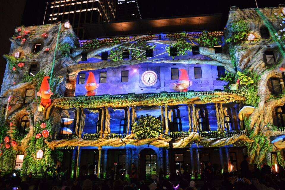 Um edifício com relógio, iluminado por luzes coloridas, cercado por vegetação e cogumelos.