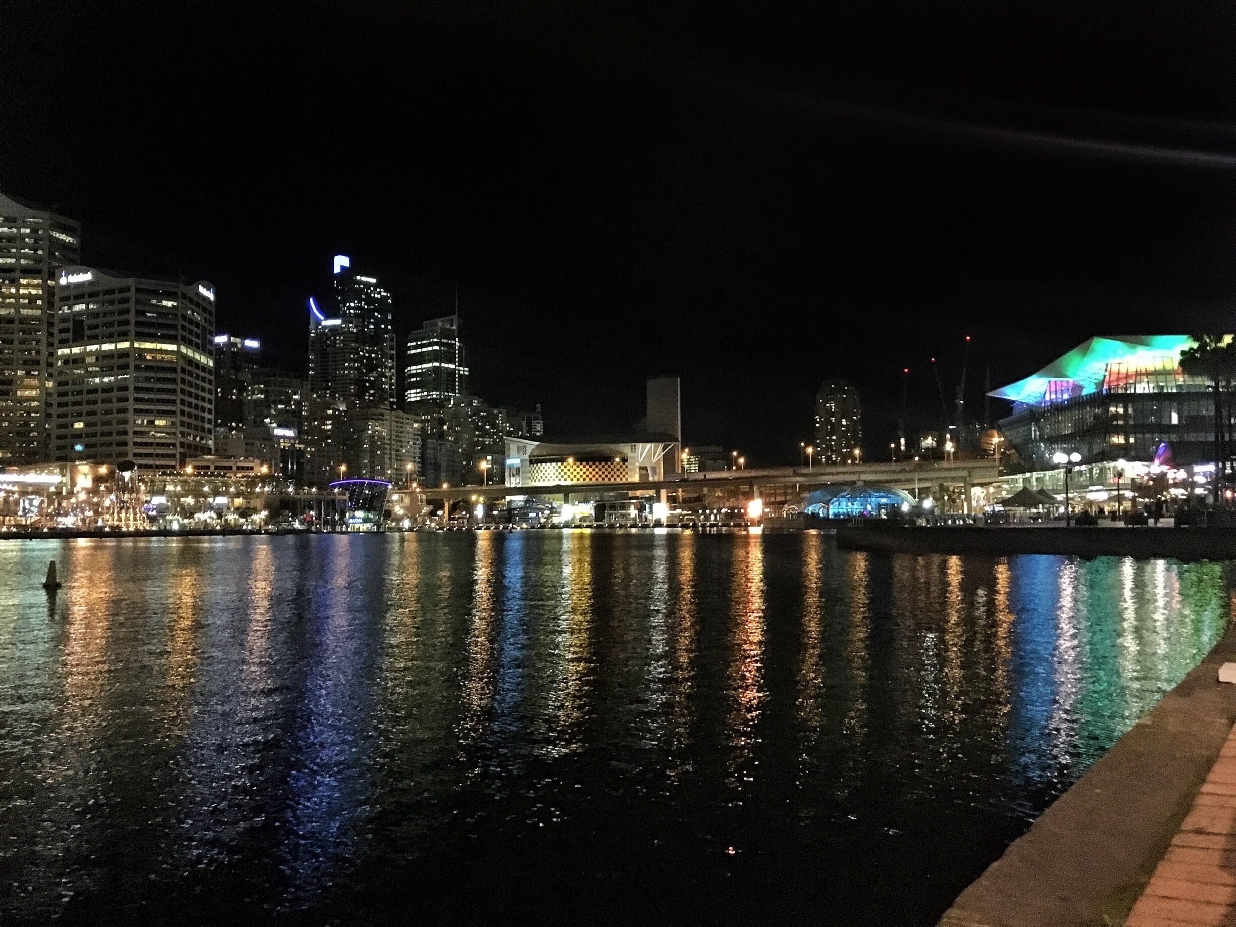 A nighttime cityscape with illuminated buildings reflecting on the water.