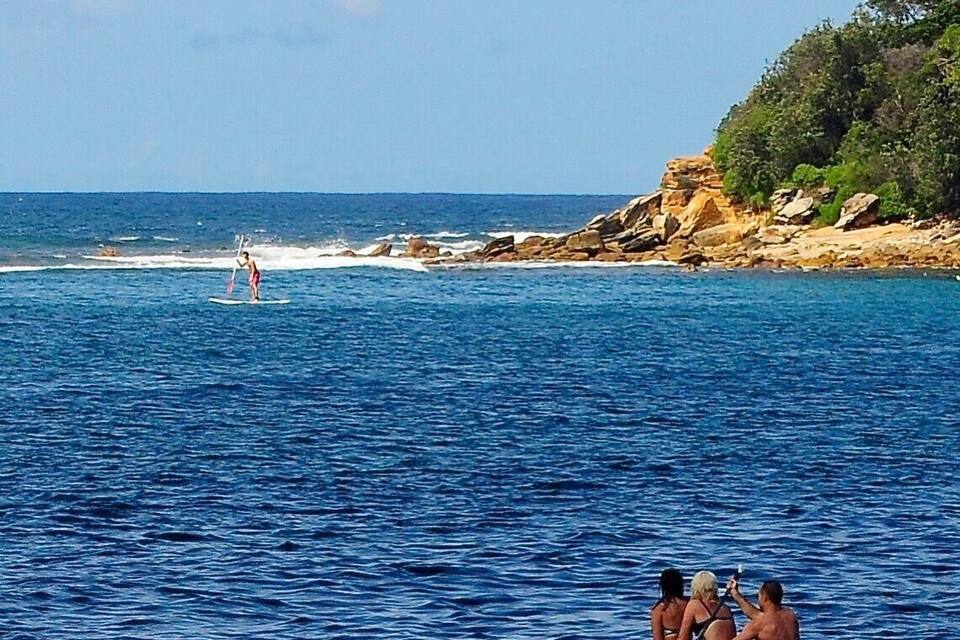 A coastal scene with a rocky shoreline, clear blue water, and a person surfing.
