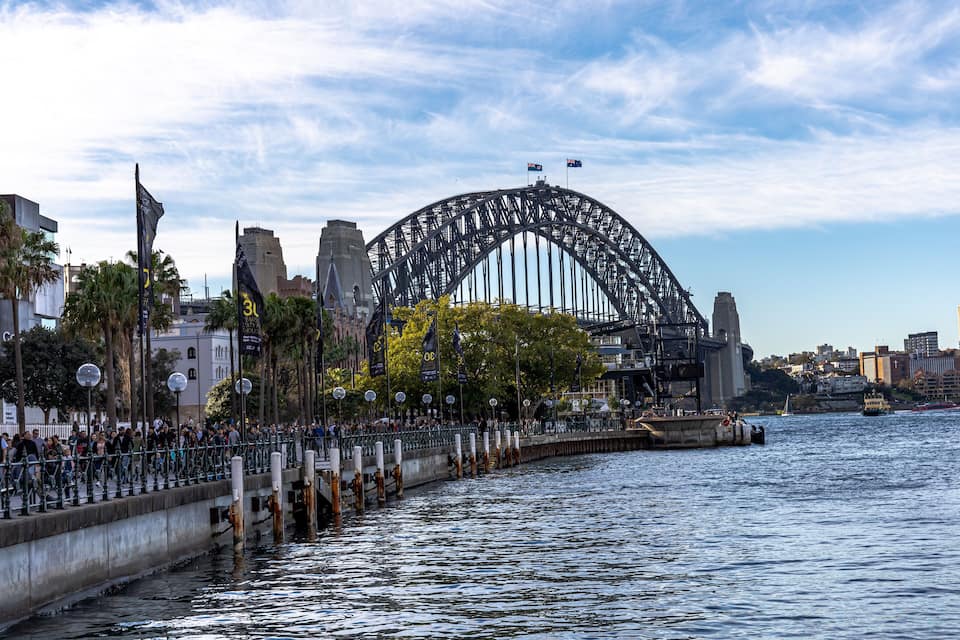 A large steel arch bridge over water with a crowd of people on the adjacent walkway.