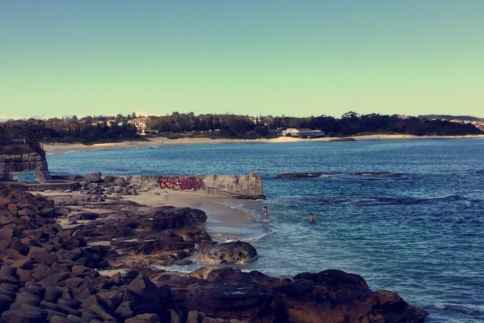 A rocky coastline with clear blue water, a sandy beach, and buildings in the background.