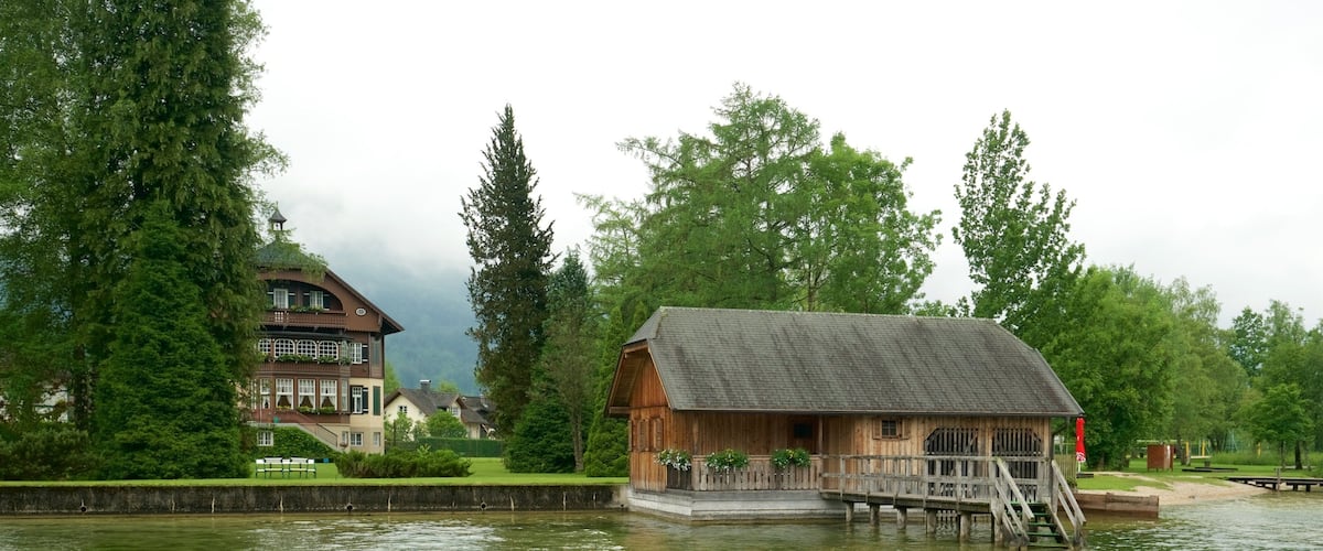 A wooden house on stilts by a calm river, surrounded by lush greenery and a large building in the background.