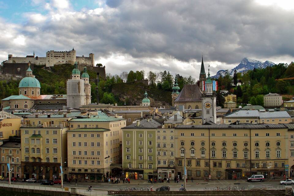 A historic cityscape with a prominent castle on a hill, a church with a green dome, and a clock tower.