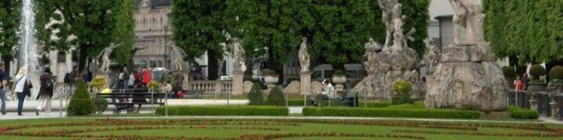A historic cityscape with a prominent castle on a hill, a fountain, and classical statues in a garden.