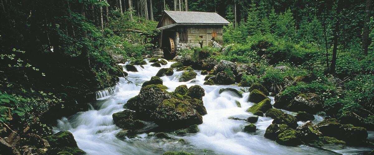 A waterfall flowing over rocks in a forest with a small cabin in the background.