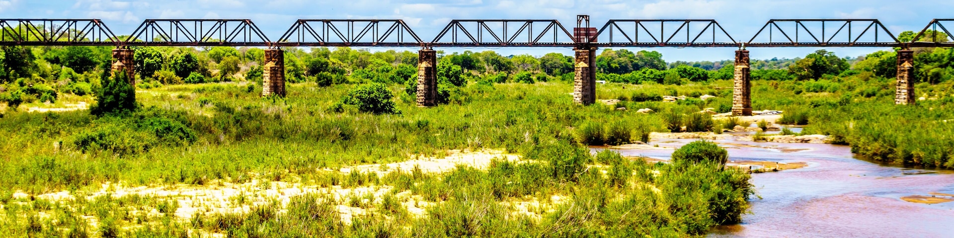 Um rio serpenteando sob uma ponte de metal com pilares de pedra, rodeado por vegetação exuberante e céu parcialmente nublado.