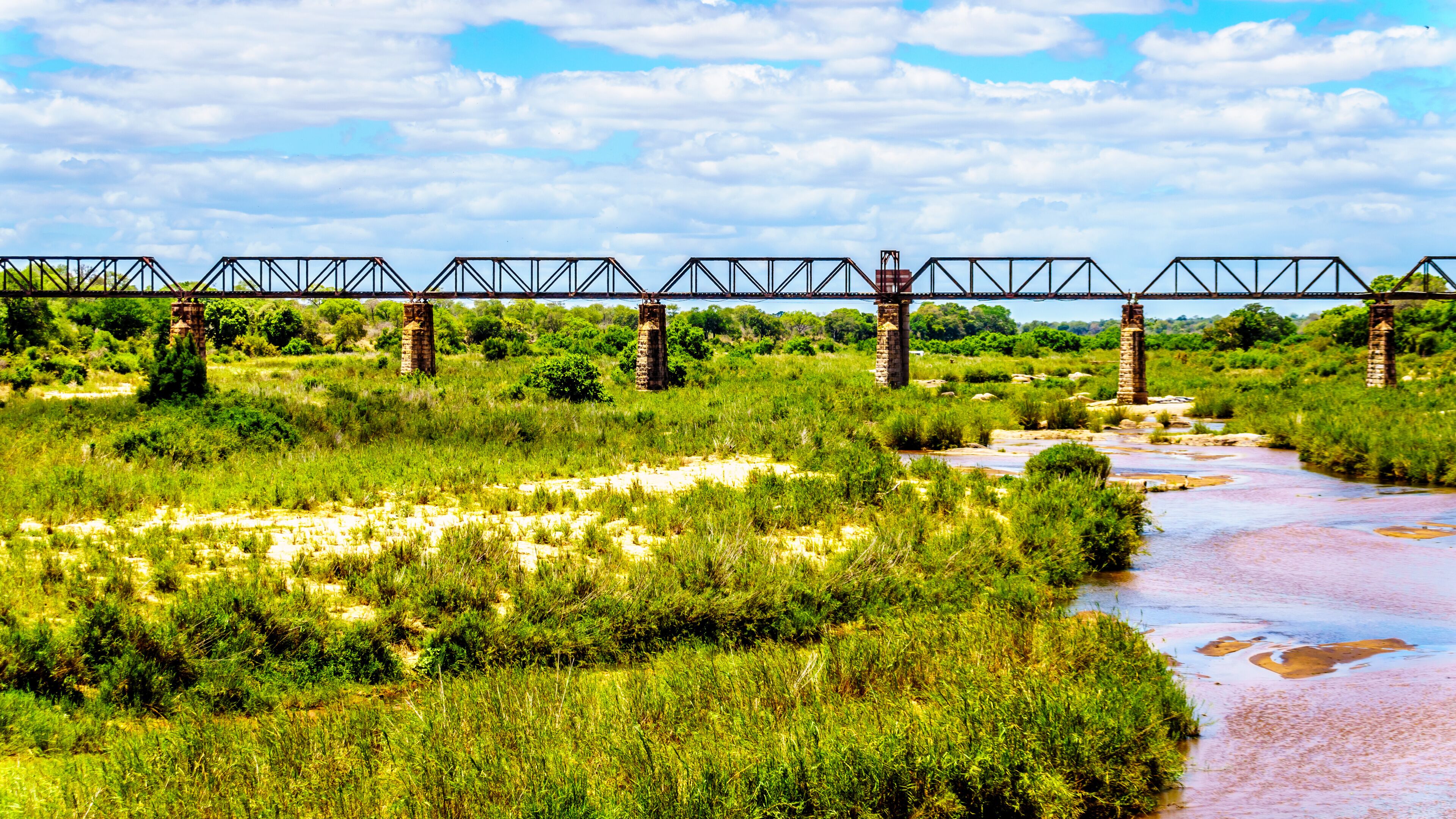 A river flowing under a metal bridge with stone pillars, surrounded by lush greenery and a partly cloudy sky.