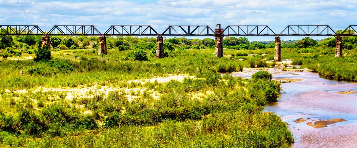A river flowing under a metal bridge with stone pillars, surrounded by lush greenery and a partly cloudy sky.