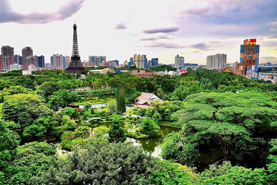 A cityscape with a prominent tower resembling the Eiffel Tower, surrounded by lush greenery and a body of water.