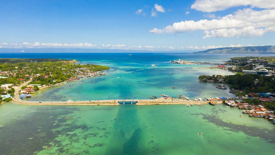 An aerial view of a coastal town with a bridge, clear turquoise waters, and a distant mountain range.