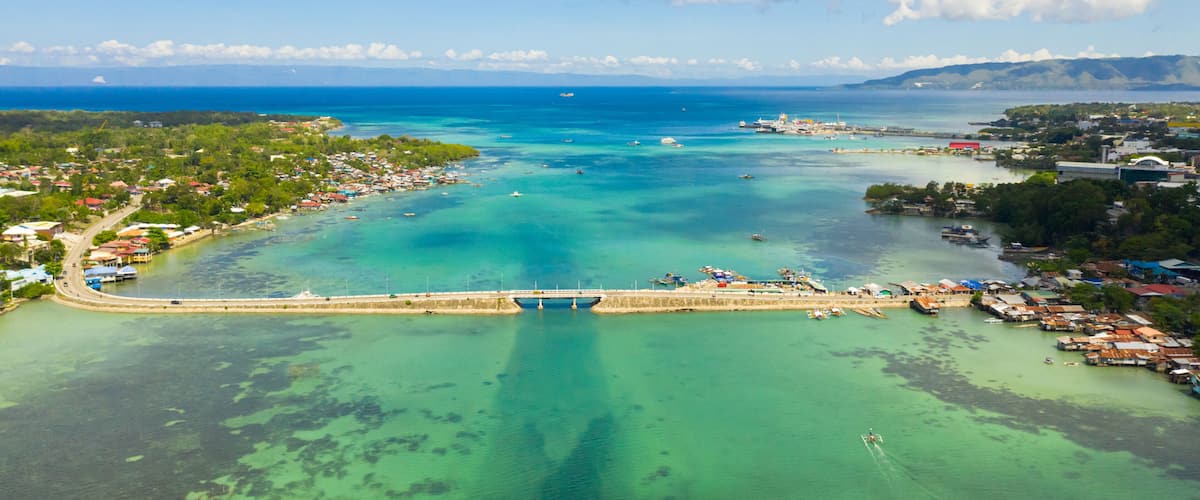 An aerial view of a coastal town with a bridge, clear turquoise waters, and a distant mountain range.