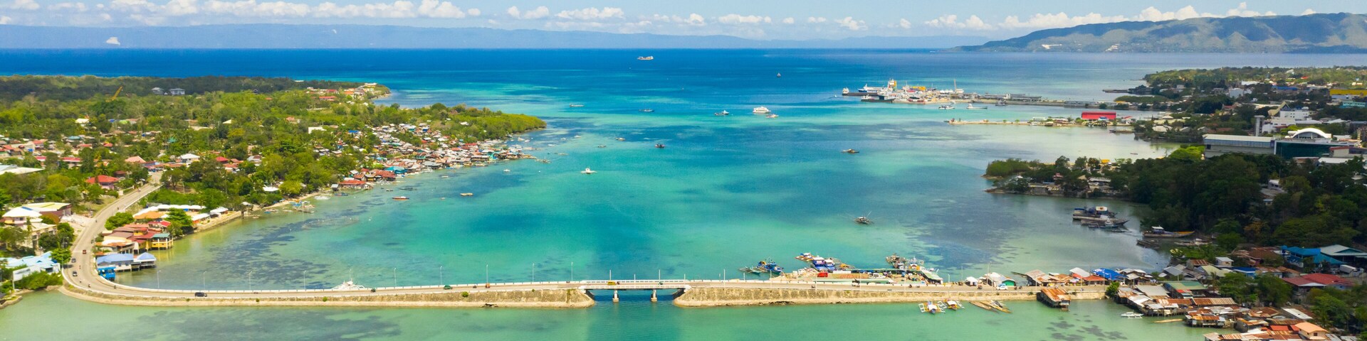 An aerial view of a coastal town with a bridge, clear turquoise waters, and a distant mountain range.