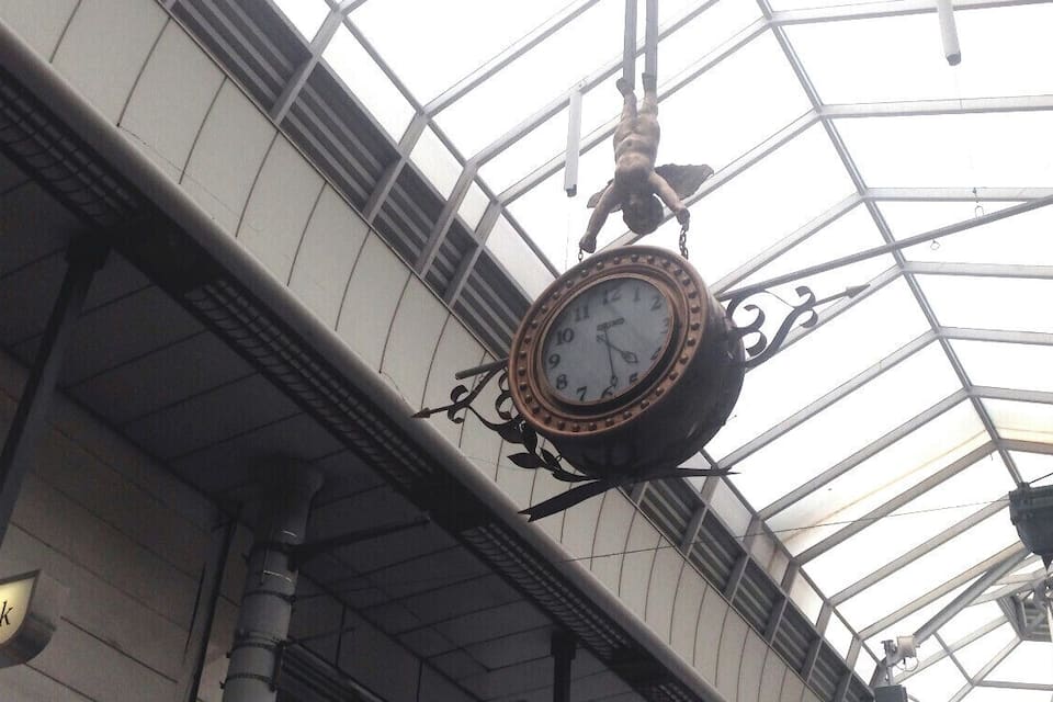 A clock hanging from a metal structure with a glass ceiling above.