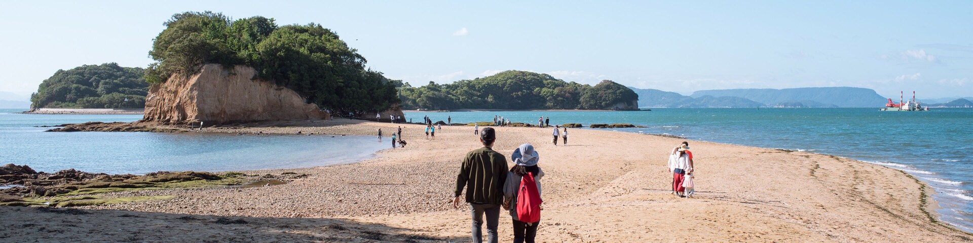 Uma praia arenosa com pessoas caminhando, uma ilha verde grande e águas azuis calmas.