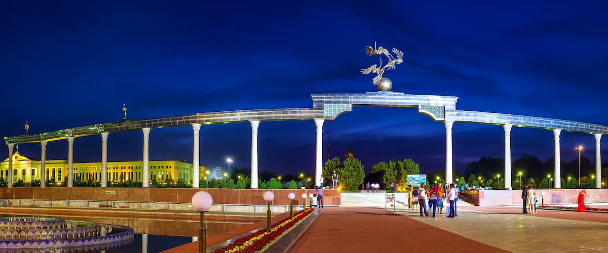 A large open-air structure with columns and a central statue, surrounded by a well-lit plaza with people and a fountain.