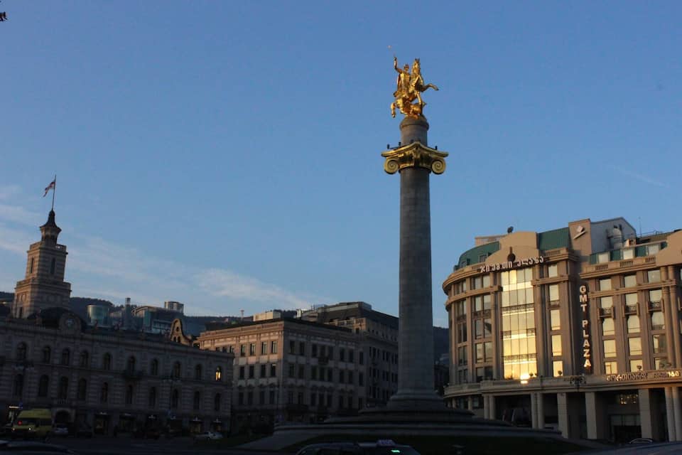 A tall monument with a golden statue, surrounded by historic buildings and a flagpole.