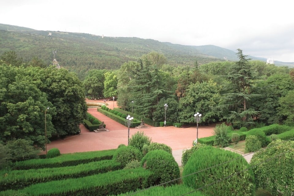 Un parque verde y exuberante con setos recortados, un camino rojo y diversas especies de árboles.