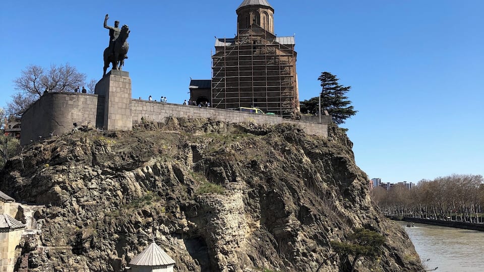 A historic fortress with a statue on top, a church with scaffolding, and a river in the foreground.