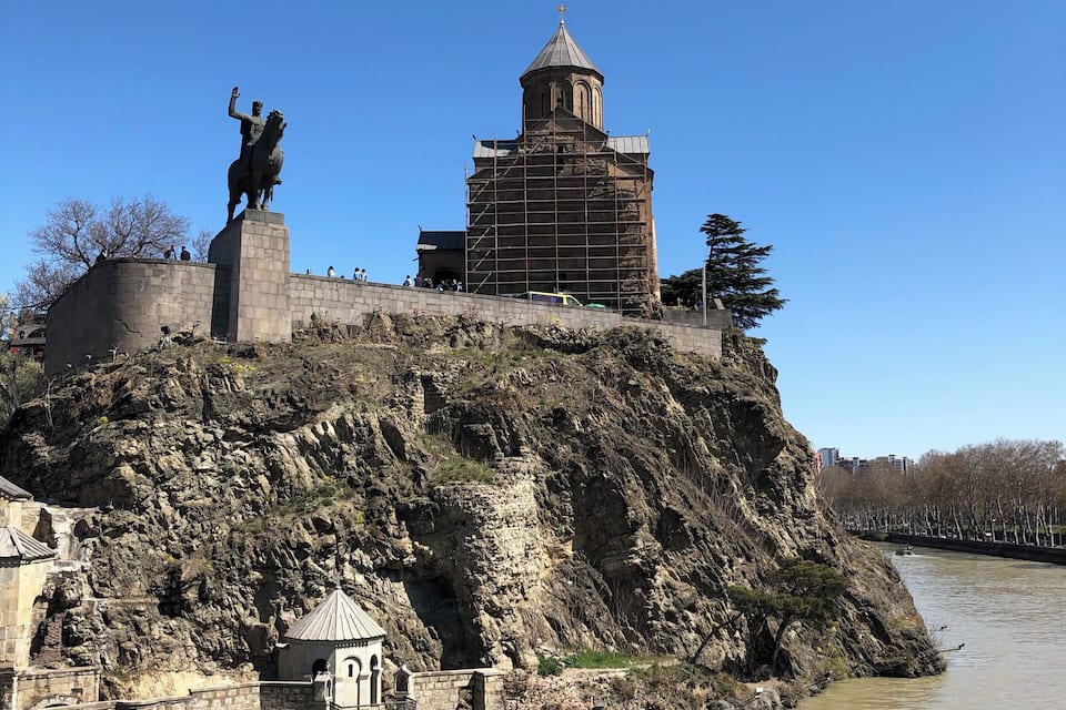 Una fortalesa històrica amb una estàtua al cim, una església amb dimec i un riu en primer pla.