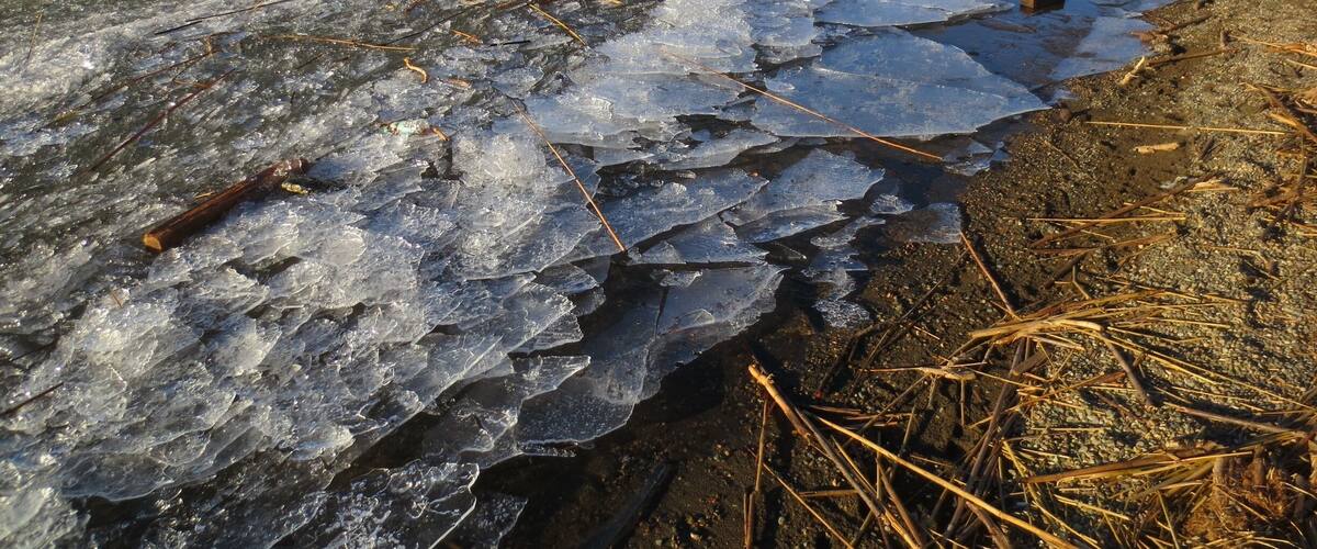 A frozen river with ice breaking, surrounded by hills and sparse vegetation.