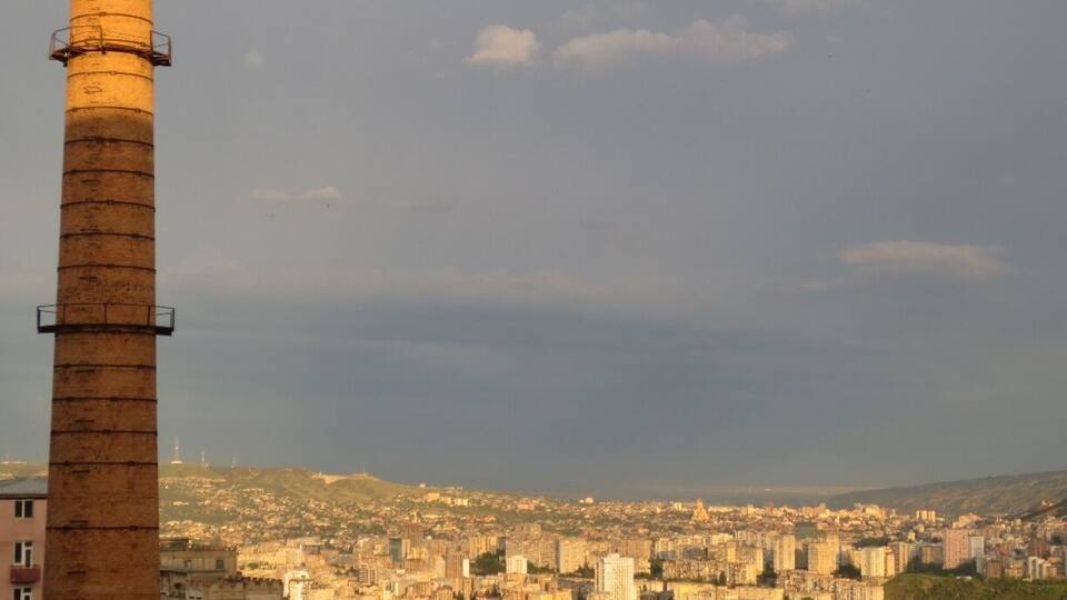 A cityscape with a prominent smokestack and a distant view of buildings and hills.