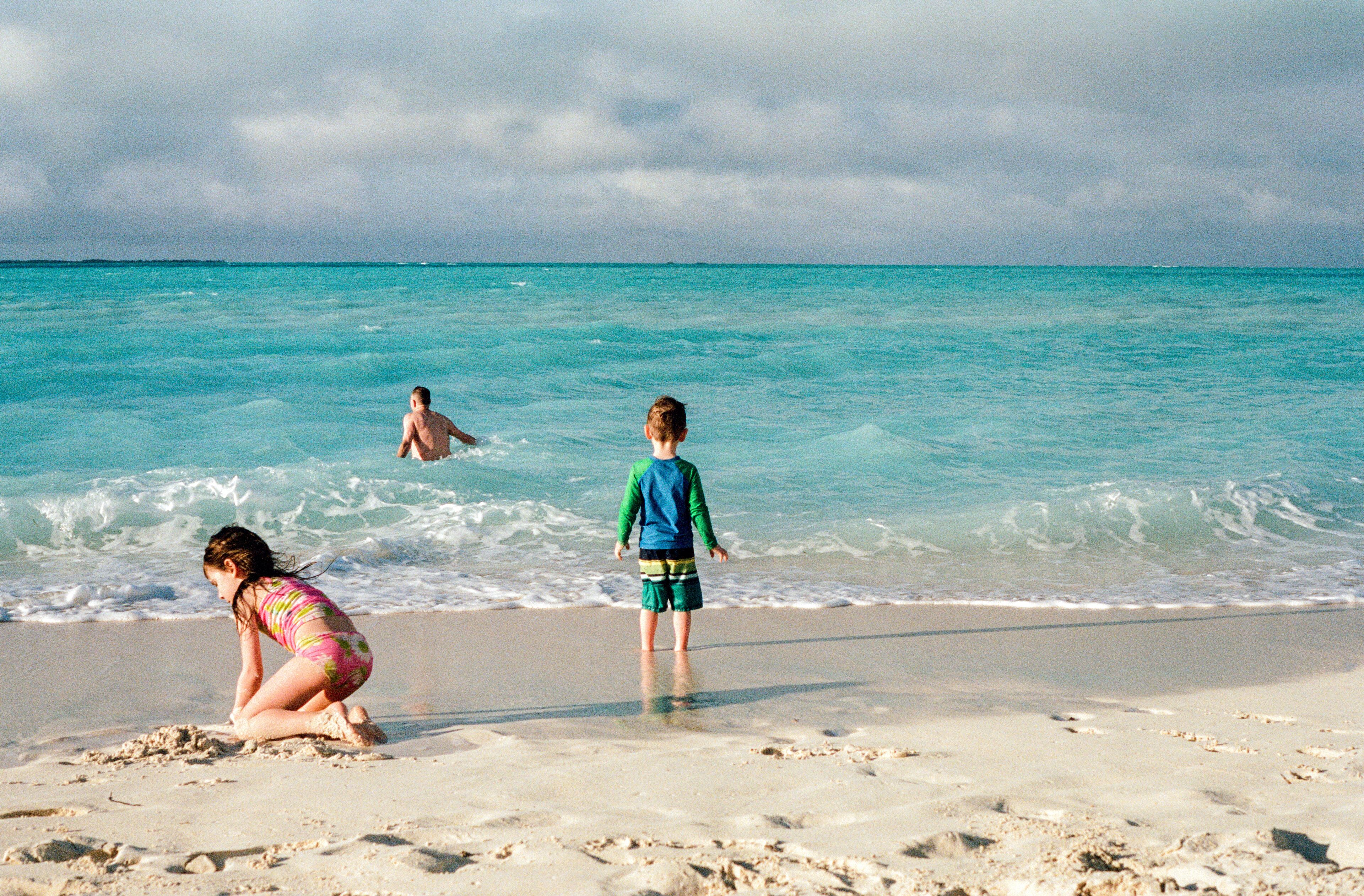 A beach with turquoise water, a child in a blue shirt, and a woman in a pink swimsuit.