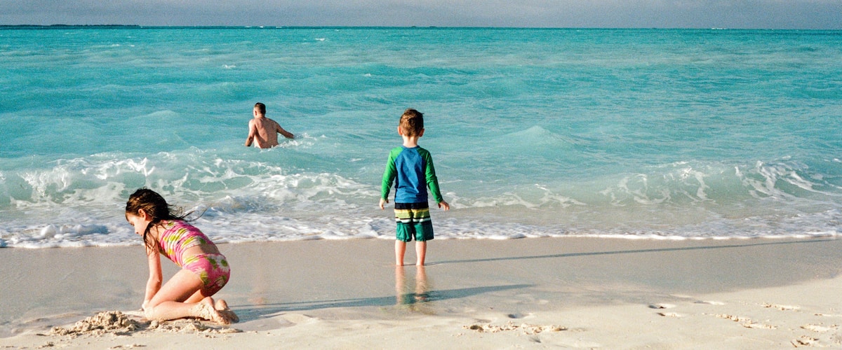 A beach with turquoise water, a child in a blue shirt, and a woman in a pink swimsuit.