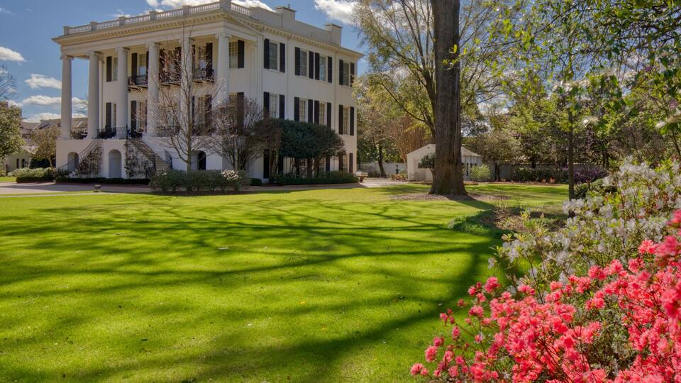 A large white building with columns, surrounded by a well-manicured lawn and flowering bushes.