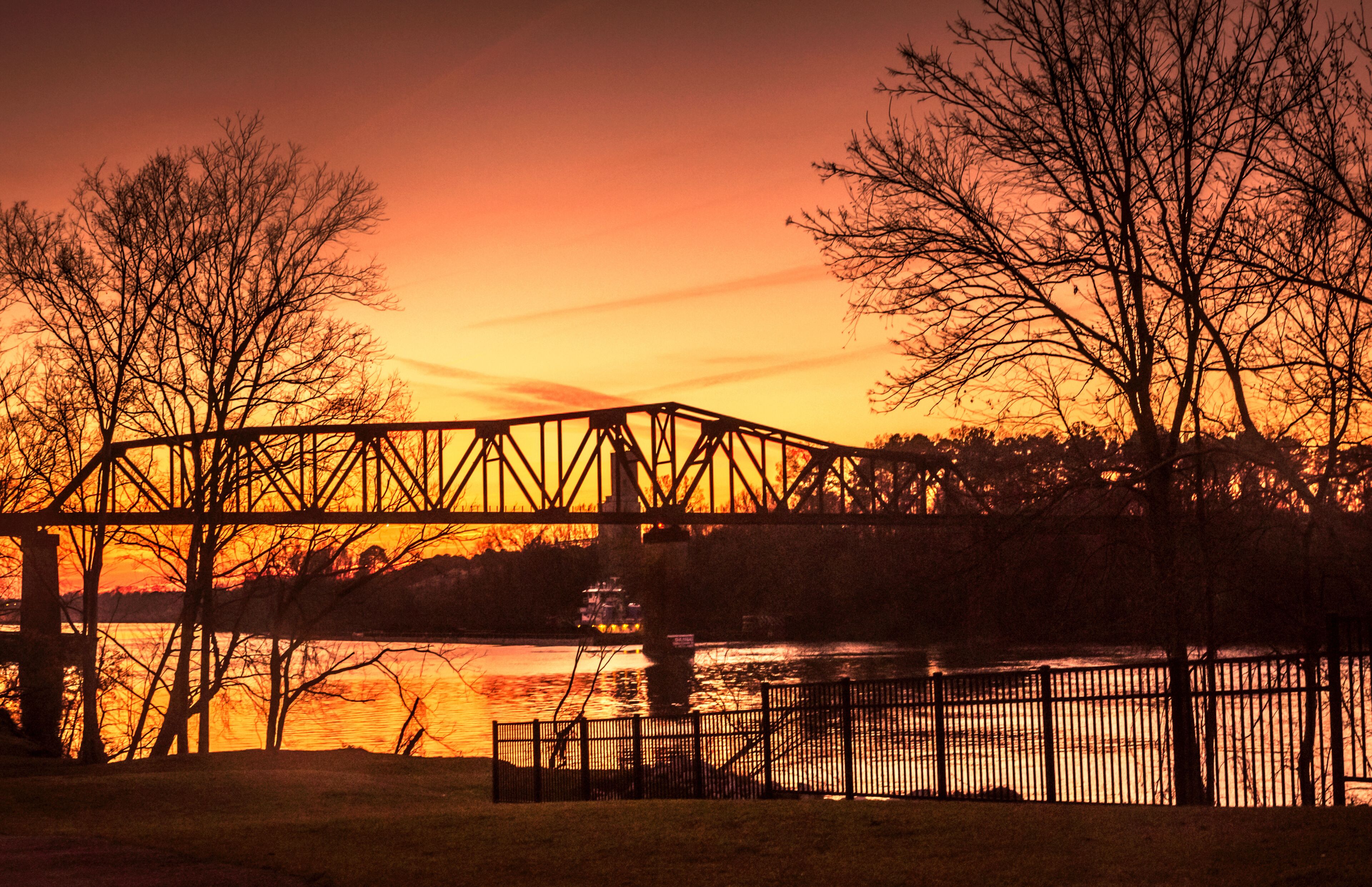 A steel bridge over a river at sunset with bare trees and a fence in the foreground.