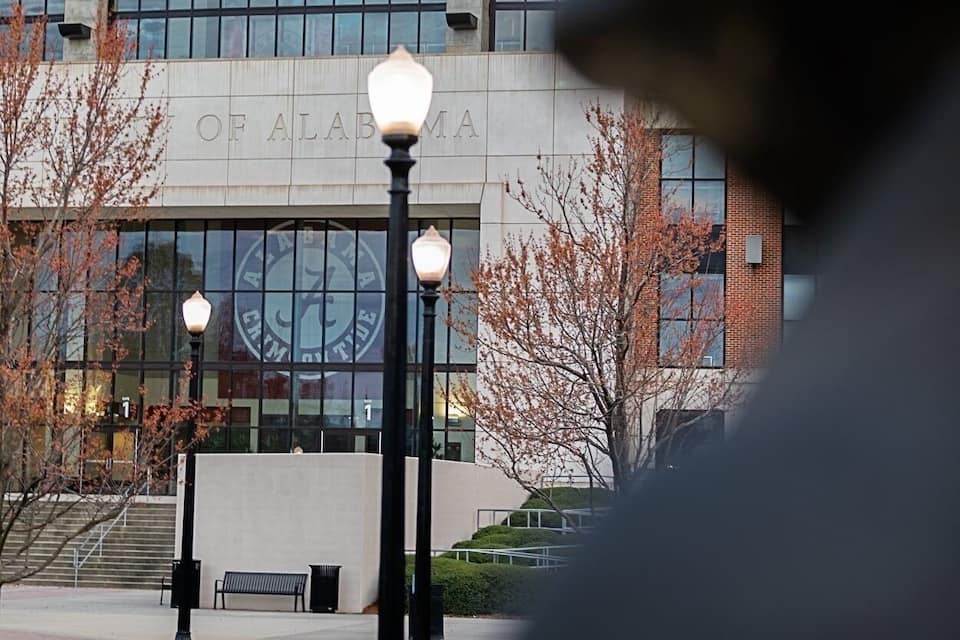 A building with a sign that reads "OF ALABAMA" and a large window. There are street lamps and a bench in the foreground.
