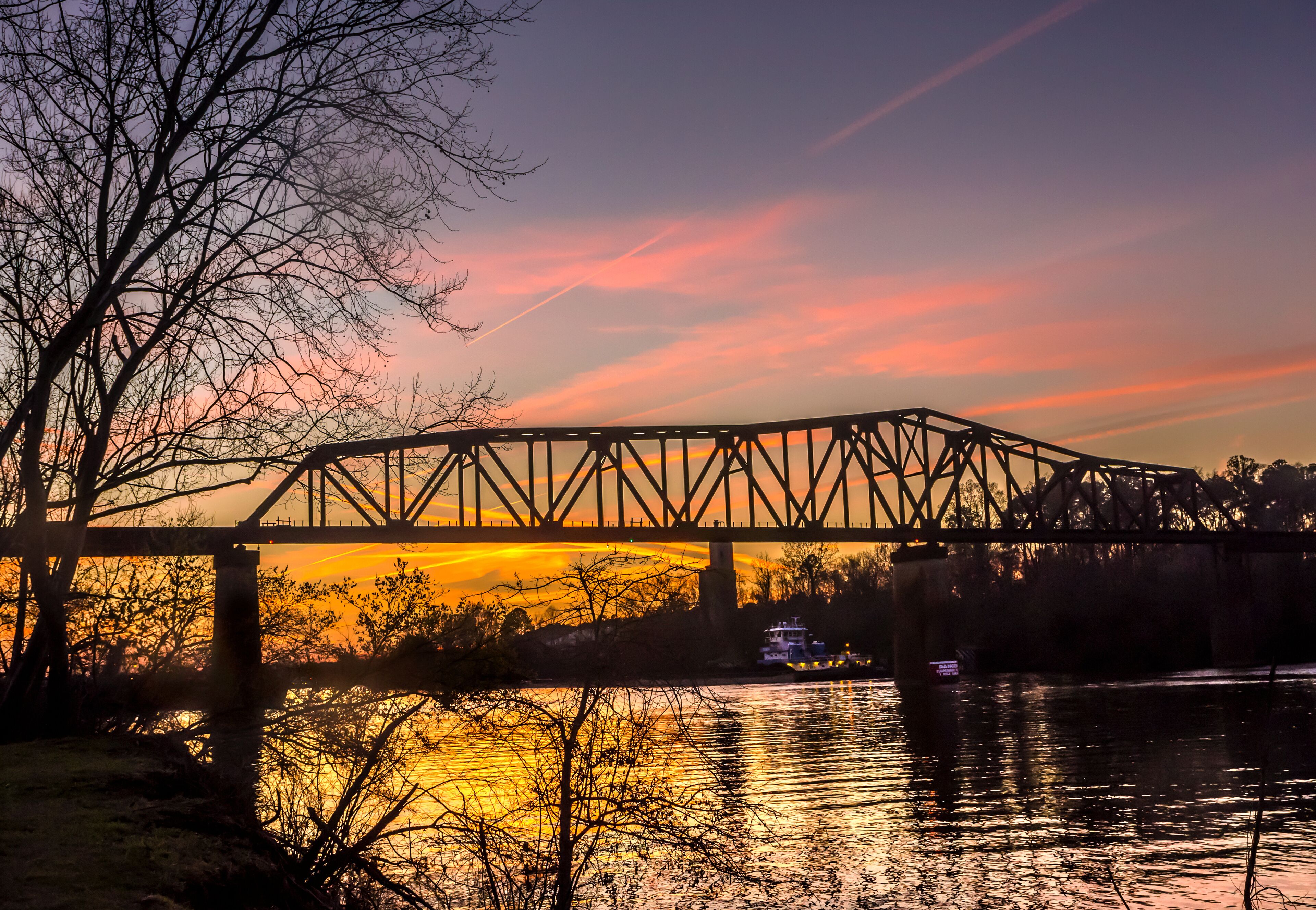 A steel bridge over a river at sunset with a reflection of the sky.