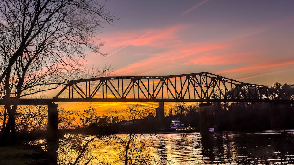 A steel bridge over a river at sunset with a reflection of the sky.