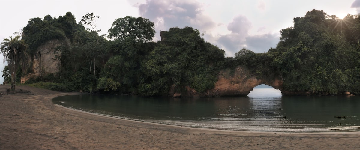 A beach with a natural rock arch formation surrounded by dense forest.
