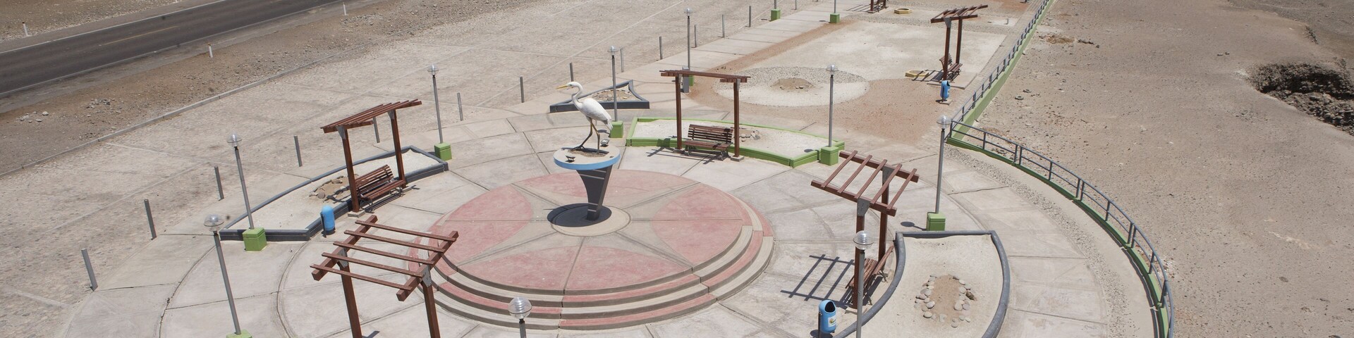 Una plaza circular con bancos y una fuente en el centro, rodeada por una carretera y montañas al fondo.