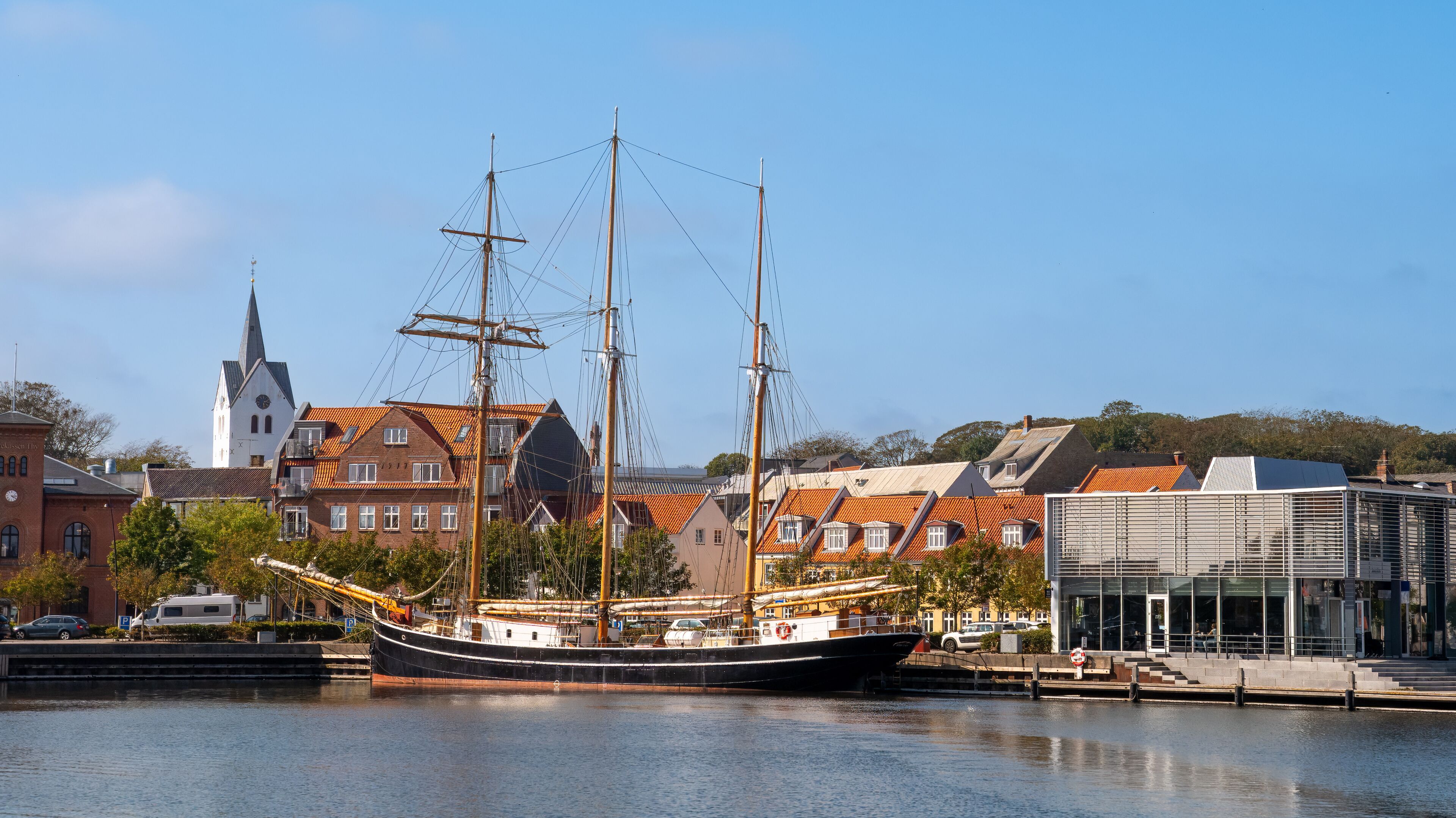 A historic sailing ship docked at a harbor with traditional buildings and a church in the background.