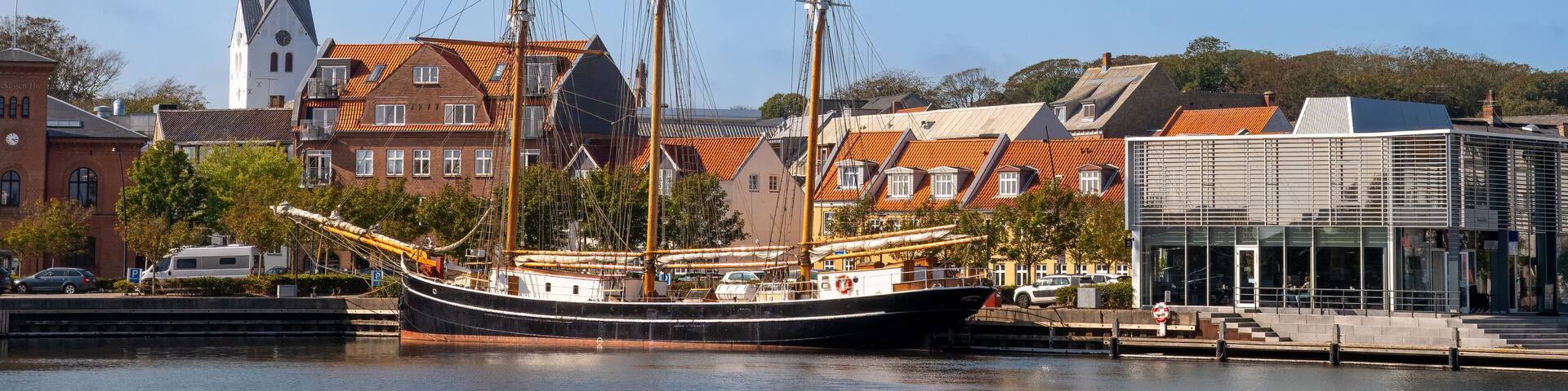 A historic sailing ship docked at a harbor with traditional buildings and a church in the background.