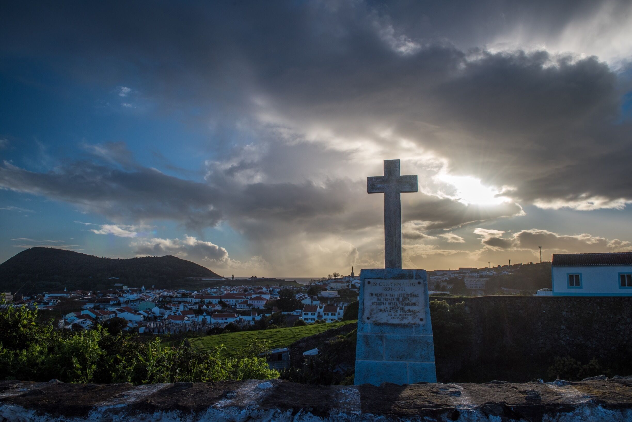 A cross monument with inscriptions, set against a backdrop of a coastal town and mountains under a partly cloudy sky.