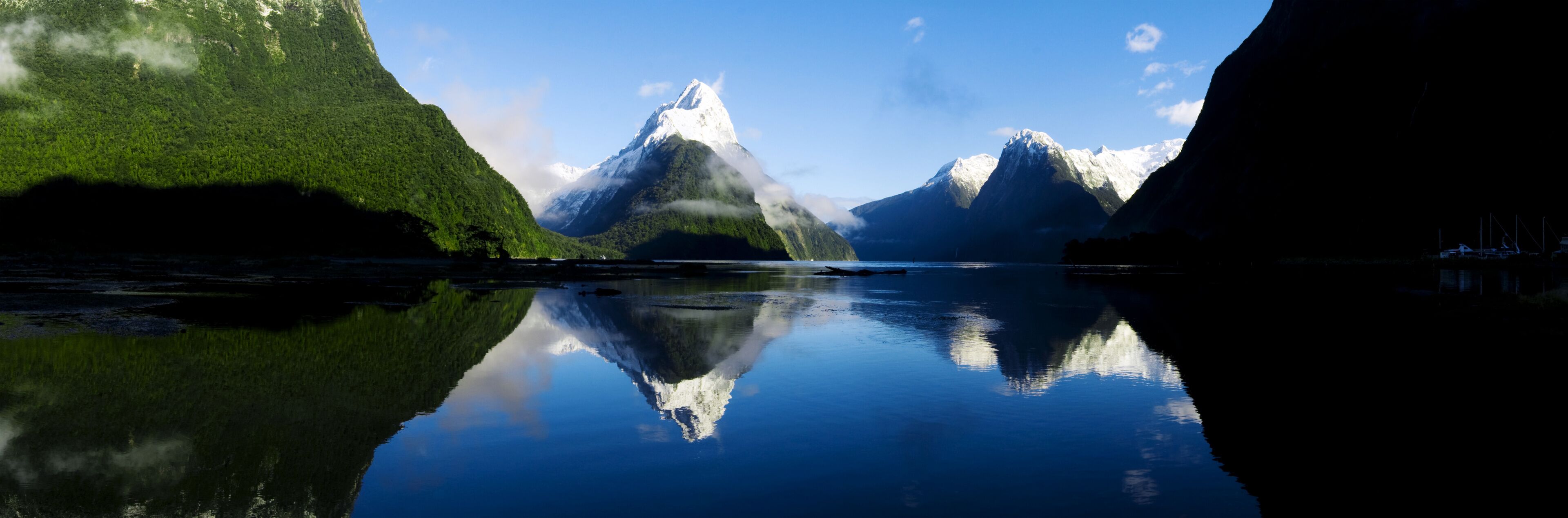 Um lago tranquilo refletindo uma cordilheira com encostas verdejantes e picos nevados.