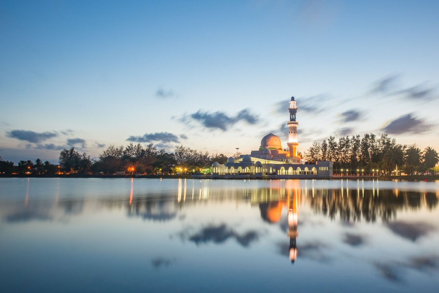Une mosquée dotée d’un minaret et d’un dôme, qui se reflète sur une étendue d’eau calme, entourée d’arbres et sous un ciel dégagé.