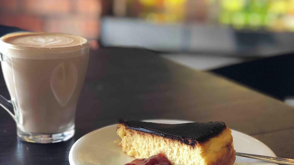 Une part de gâteau à la surface foncée, présentée sur une assiette blanche, accompagnée d’une tasse de café ornée d’un motif en latte art.