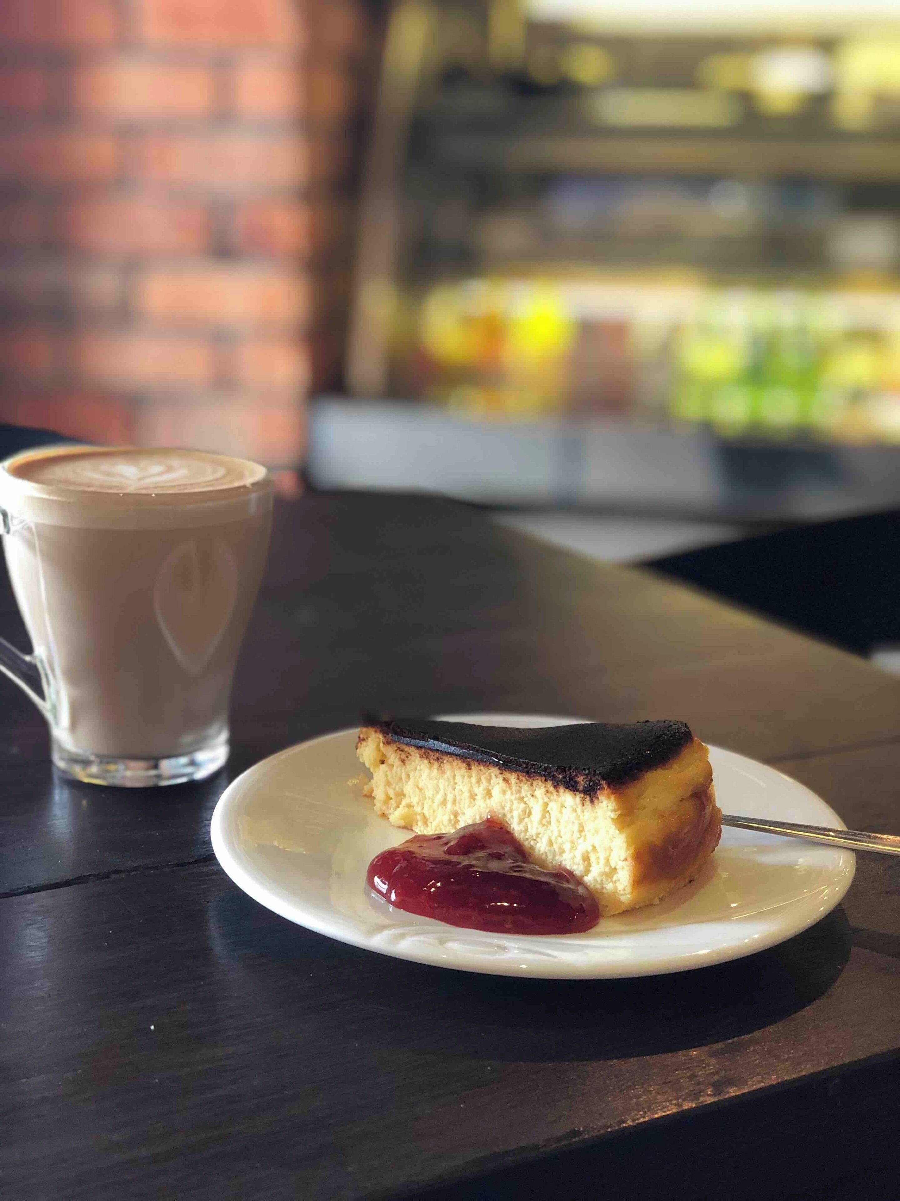 A slice of cake with a dark top on a white plate, accompanied by a cup of coffee with latte art.