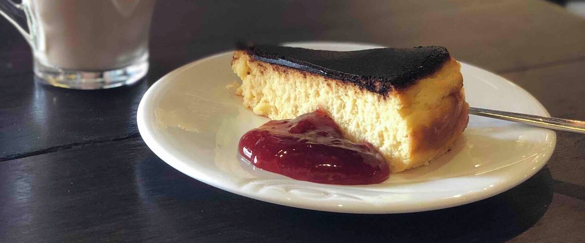 A slice of cake with a dark top on a white plate, accompanied by a cup of coffee with latte art.